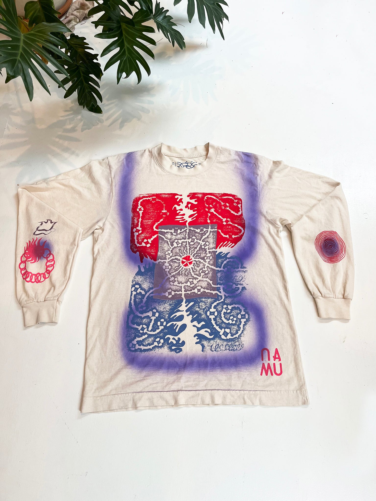 NAMU's first t0shirt collaboration with artist Maren Jensen, a whiite long-sleeve cotton tshirt screenprinted with a multi color maze-like graphic overlayed with a soft purple airbrushed shape lays spread out against a white background under a green plant.