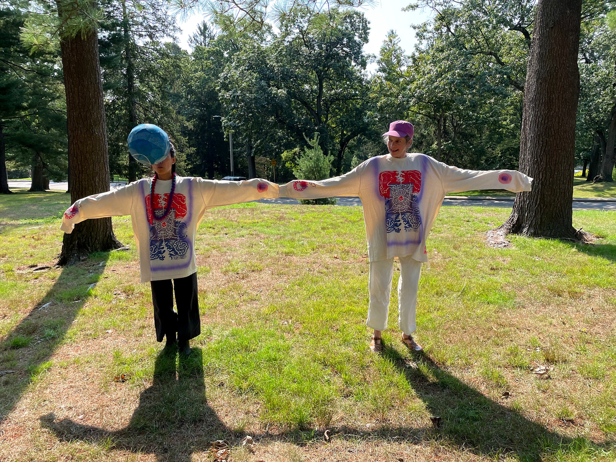 Jung and Kate stand on grass between two trees wearing Maren Jensen's Maze Tee with their arms spread in the sun.